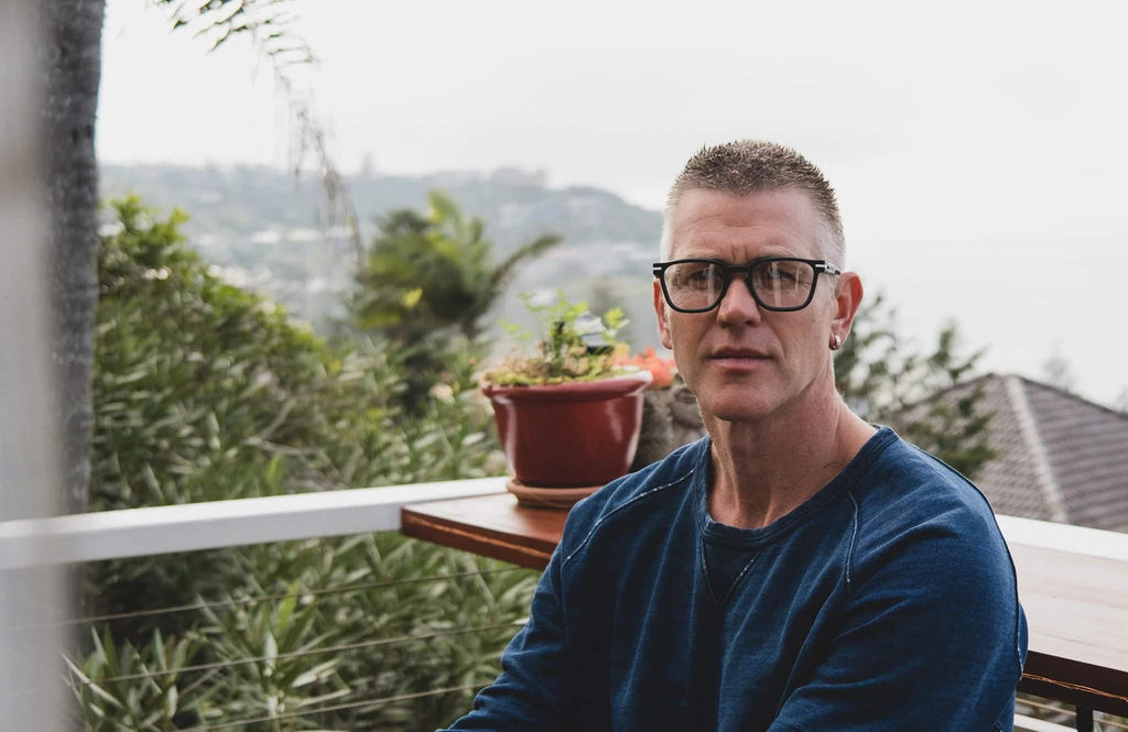 Man wearing Good Citizens black prescription glasses on a balcony with coastal views, made in Sydney from 100 percent recycled frames.