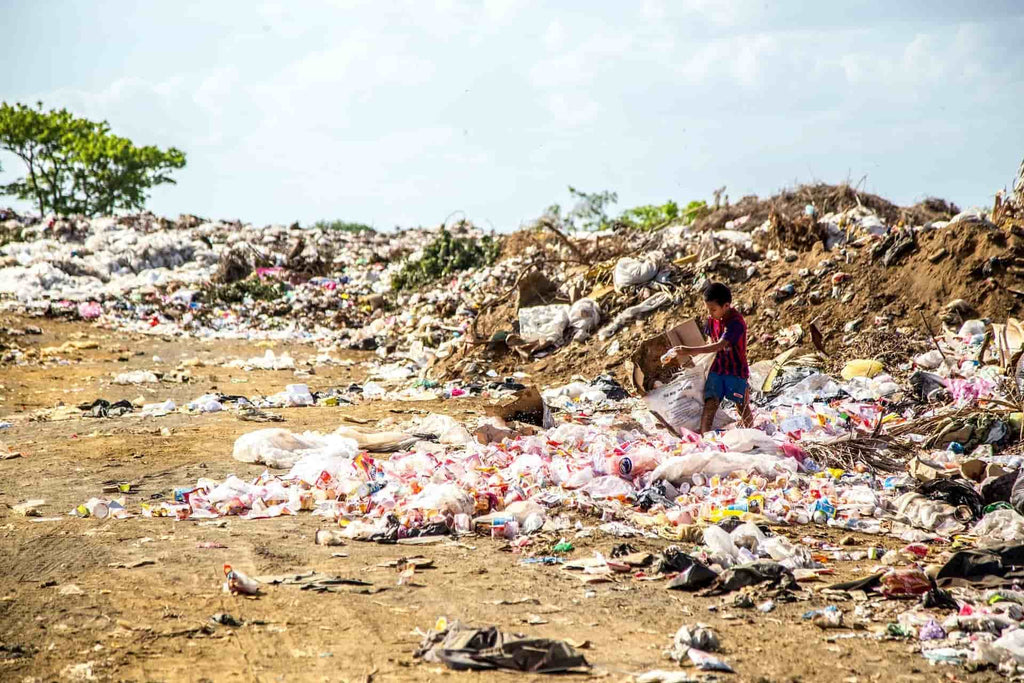 Child collecting rubbish in a large landfill under the hot sun with piles of plastic waste.